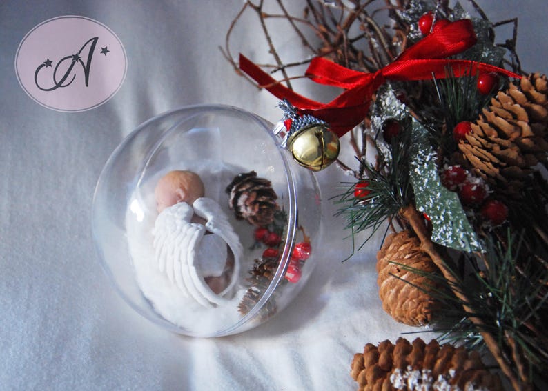 May include: A clear glass ornament with a small, white porcelain angel inside. The ornament is surrounded by pine branches, pine cones, and red berries.