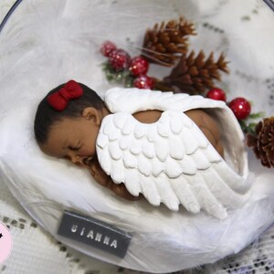 May include: A white porcelain angel figurine with wings, wearing a red bow and sleeping on a bed of white cotton. The figurine is inside a clear glass dome with a brown pine cone and red berries.