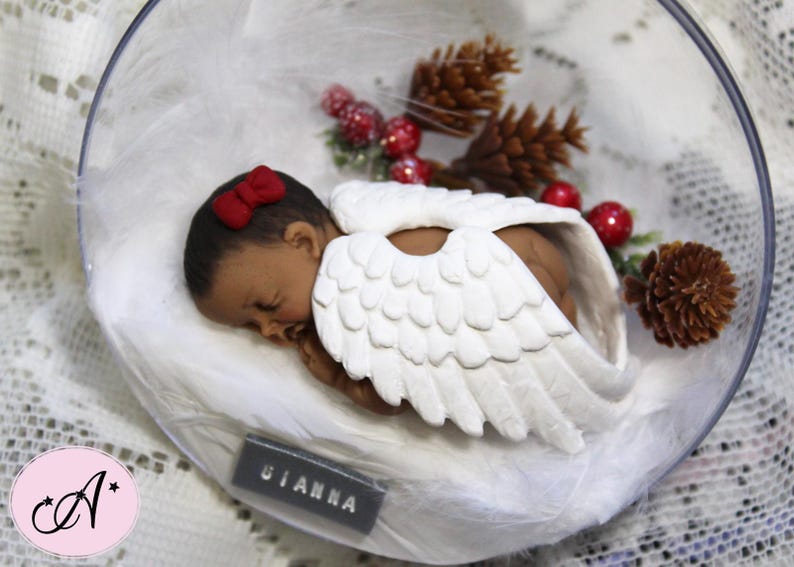 May include: A white porcelain angel figurine with wings, wearing a red bow and sleeping on a bed of white cotton. The figurine is inside a clear glass dome with a brown pine cone and red berries.