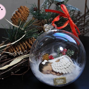 May include: A clear glass Christmas ornament with a white baby angel inside. The angel is wearing a white feathered wing and has a gold bow on its head. The ornament is decorated with white feathers and red berries. The ornament has a gold tag with the name "Sylvia Drew" on it.