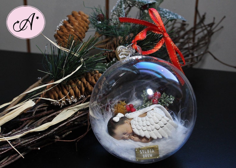 May include: A clear glass Christmas ornament with a white baby angel inside. The angel is wearing a white feathered wing and has a gold bow on its head. The ornament is decorated with white feathers and red berries. The ornament has a gold tag with the name "Sylvia Drew" on it.