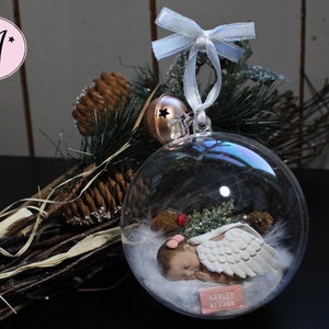 May include: A clear glass ornament with a sleeping baby angel inside. The angel has white wings and is lying on a bed of white feathers. The ornament is decorated with a small red bow and the name "Harley Alydia" is written on a tag.