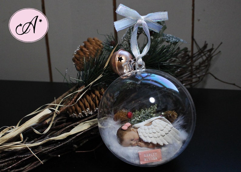 May include: A clear glass ornament with a sleeping baby angel inside. The angel has white wings and is lying on a bed of white feathers. The ornament is decorated with a small red bow and the name "Harley Alydia" is written on a tag.