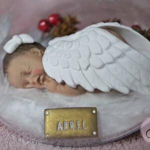 May include: A white porcelain figurine of a sleeping baby angel with wings. The baby is wearing a white bow and is lying on a bed of white feathers. A gold tag with the name "APRIL" is in the foreground.