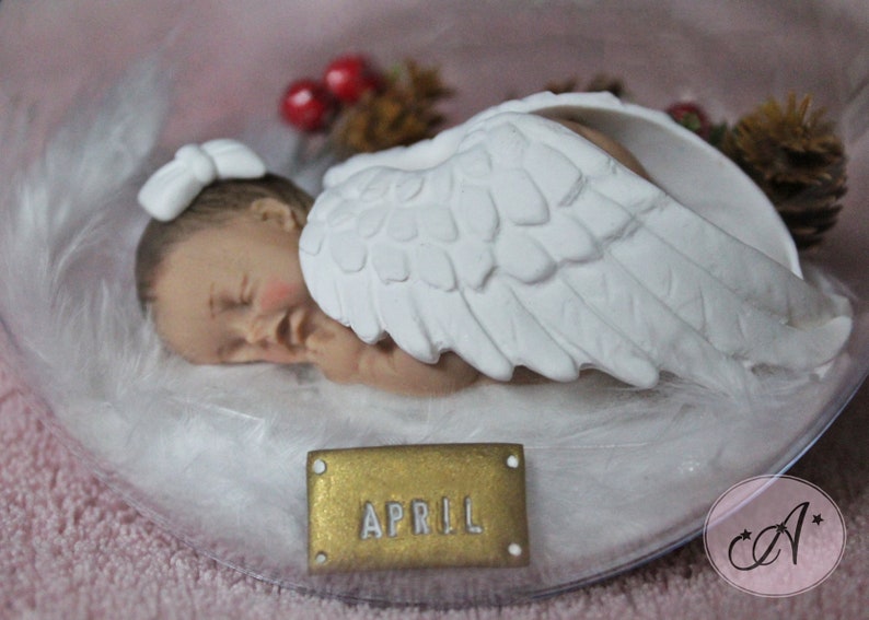 May include: A white porcelain figurine of a sleeping baby angel with wings. The baby is wearing a white bow and is lying on a bed of white feathers. A gold tag with the name "APRIL" is in the foreground.
