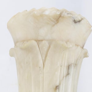 May include: A carved alabaster vase with a flared top and a pedestal base. The vase is off-white with subtle brown veining and a square base. The design features vertical grooves and a floral-like top.