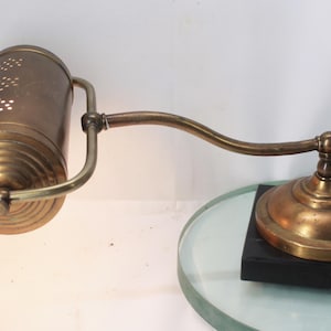 May include: A vintage brass desk lamp with a cylindrical shade featuring decorative perforations. The lamp has an adjustable arm and a round base, mounted on a black rectangular base. The lamp's shade is approximately 6 inches in diameter.
