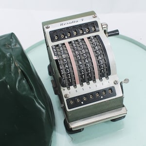 May include: A vintage Resulta 7 mechanical calculator with a green and silver finish. The calculator features multiple number dials and a handle. The text "Resulta 7" is visible on the top. A green case is on the left.