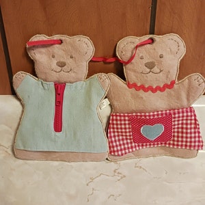 May include: Two plush teddy bear ornaments. One bear wears a light blue shirt with a red zipper, the other a red and white checkered apron with a blue heart. Both have red ribbon loops for hanging.