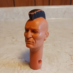 May include: A plastic action figure head with a mohawk hairstyle. The head is light brown and has a detailed face with a serious expression. The mohawk is black and is styled in a spiky fashion.