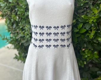 May include: White sleeveless dress with a scoop neckline. The dress features a band of navy blue bow patterns around the waist. The dress has a flared skirt and is made of a knitted fabric.