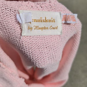 May include: A light pink knitted garment with a tag that reads "tennisknit by Hampton Court." The tag also includes washing instructions. The garment is made of a soft, textured material.