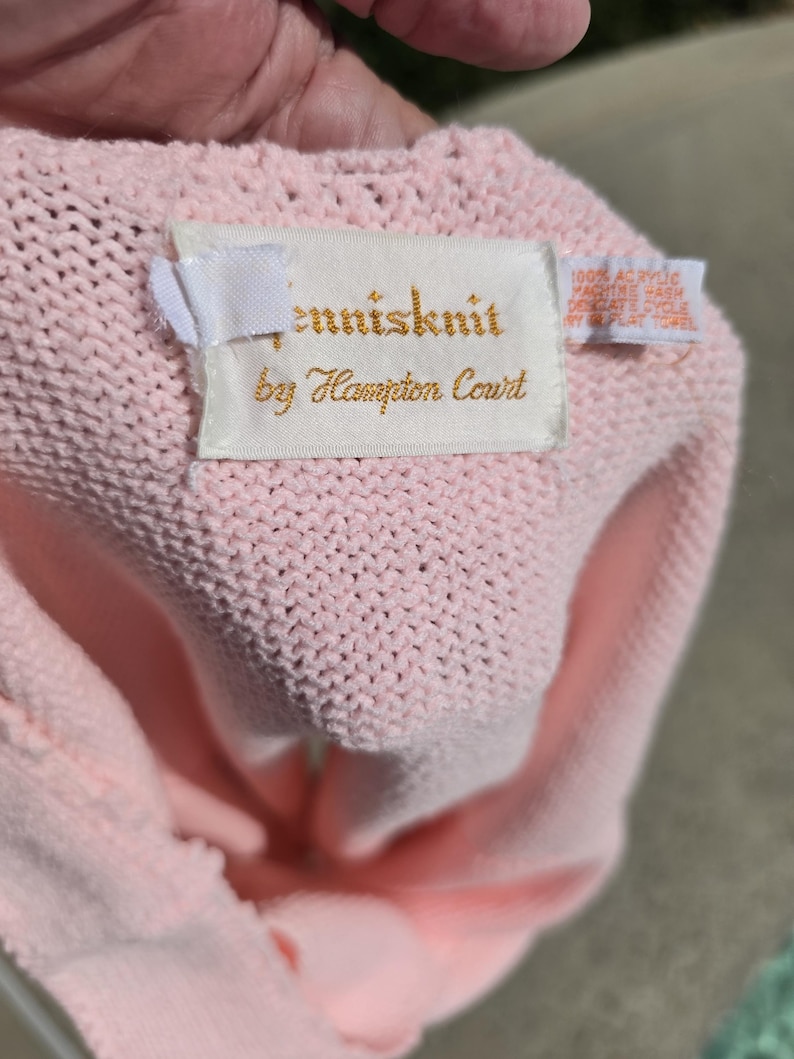 May include: A light pink knitted garment with a tag that reads "tennisknit by Hampton Court." The tag also includes washing instructions. The garment is made of a soft, textured material.