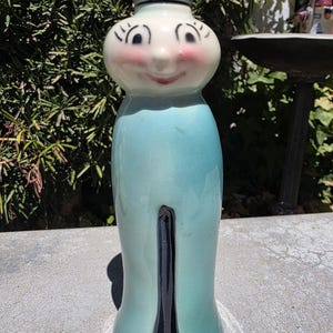 May include: A ceramic shaker featuring a stylised figure with a round, cream-coloured head, painted facial features including eyelashes and rosy cheeks, and a smooth, light blue body with a vertical black stripe extending from the base upwards.