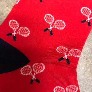 May include: Red socks patterned with white tennis rackets with black handles. The heel and toe are navy blue. These socks are ideal for anyone who enjoys playing tennis.