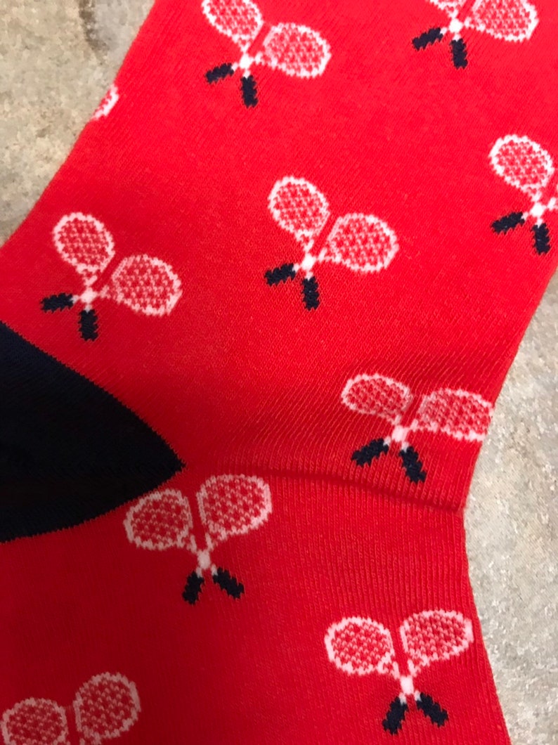 May include: Red socks patterned with white tennis rackets with black handles. The heel and toe are navy blue. These socks are ideal for anyone who enjoys playing tennis.