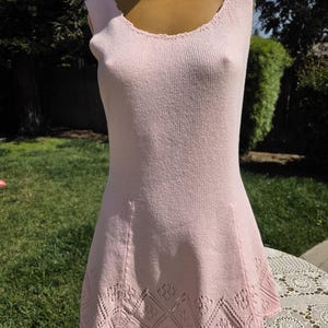 May include: A light pink sleeveless dress with a scoop neckline and a flared skirt. The dress is made of a knitted fabric and features a decorative diamond pattern near the hemline. The dress is displayed on a mannequin outdoors.