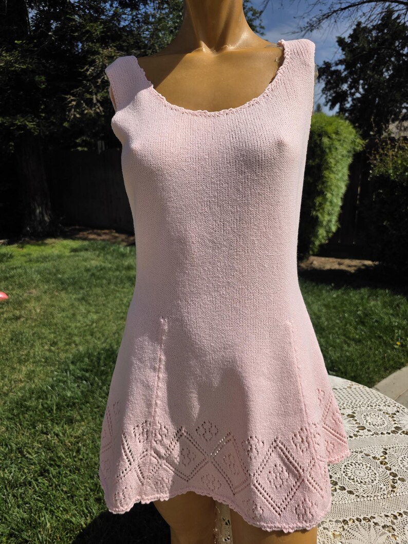 May include: A light pink sleeveless dress with a scoop neckline and a flared skirt. The dress is made of a knitted fabric and features a decorative diamond pattern near the hemline. The dress is displayed on a mannequin outdoors.