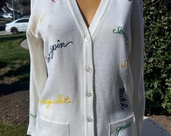 May include: White cardigan sweater with colorful embroidered words like "add", "top spin", "serve", "match", "game", "love", and "chip shot".
