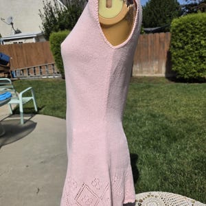 May include: A light pink sleeveless knit dress with a decorative diamond pattern at the hem. The dress is displayed on a mannequin outdoors, showcasing its textured fabric and design details. The dress is a mini dress.