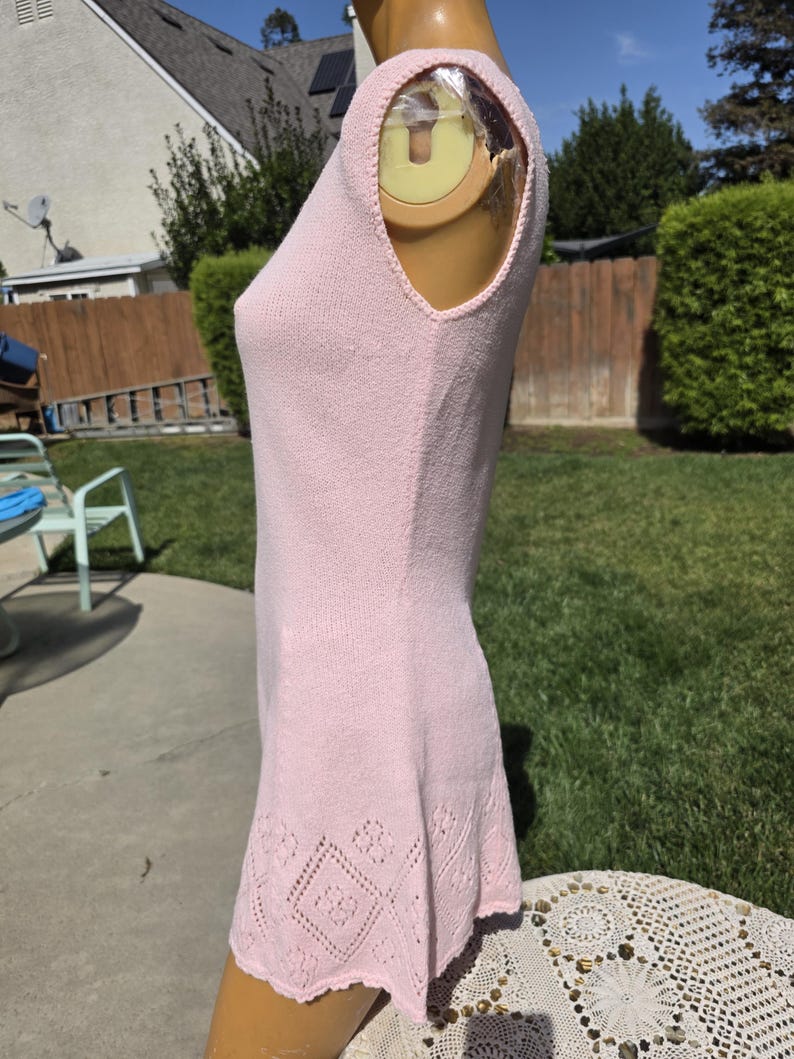 May include: A light pink sleeveless knit dress with a decorative diamond pattern at the hem. The dress is displayed on a mannequin outdoors, showcasing its textured fabric and design details. The dress is a mini dress.