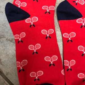 May include: A pair of red socks featuring a pattern of white tennis rackets. The heels and toes are navy blue. The tennis rackets are detailed with white strings and black handles.