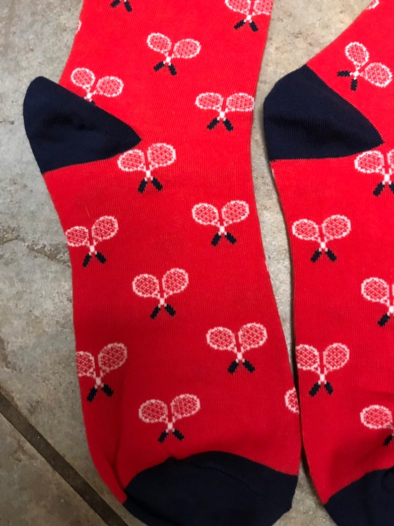 May include: A pair of red socks featuring a pattern of white tennis rackets. The heels and toes are navy blue. The tennis rackets are detailed with white strings and black handles.