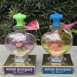 Puede incluir: Dos Kiddle Kolognes vintage de Mattel, con muñecas en miniatura dentro de botellas transparentes en forma de corazón. Uno tiene un tapón verde y una muñeca violeta, el otro un tapón azul y una muñeca naranja. Cada uno se asienta sobre una base dorada con una etiqueta.