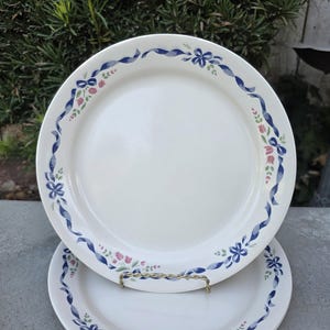 May include: Two white ceramic plates with a blue ribbon and floral design around the rim. The plates are stacked, with one resting on a gold-colored metal plate stand. The floral design includes pink and green accents.