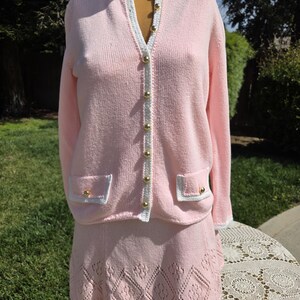 May include: A light pink knit cardigan and skirt set. The cardigan has a white trim, gold buttons, and two front pockets. The skirt is a matching light pink with a diamond pattern at the hem. The set is displayed on a mannequin.