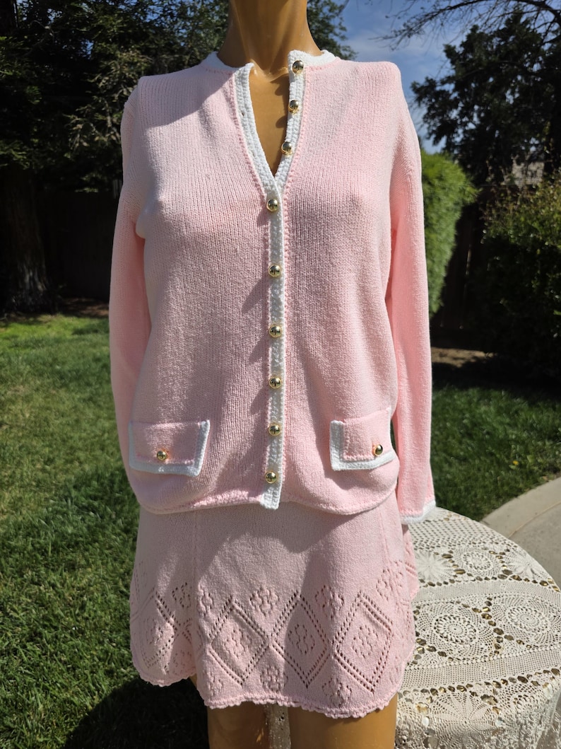 May include: A light pink knit cardigan and skirt set. The cardigan has a white trim, gold buttons, and two front pockets. The skirt is a matching light pink with a diamond pattern at the hem. The set is displayed on a mannequin.