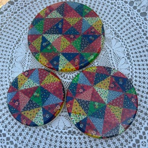 May include: Set of three colorful round trivets with a patchwork design. The trivets are made of a hard, heat-resistant material and feature a variety of colors, including red, blue, green, yellow, and white.