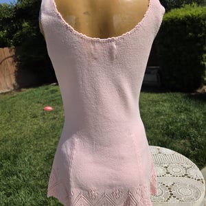 May include: A light pink sleeveless knit dress. The dress features a scoop neckline and a decorative openwork hemline. The dress is displayed on a mannequin outdoors, with a green lawn and a white lace-covered table in the background.