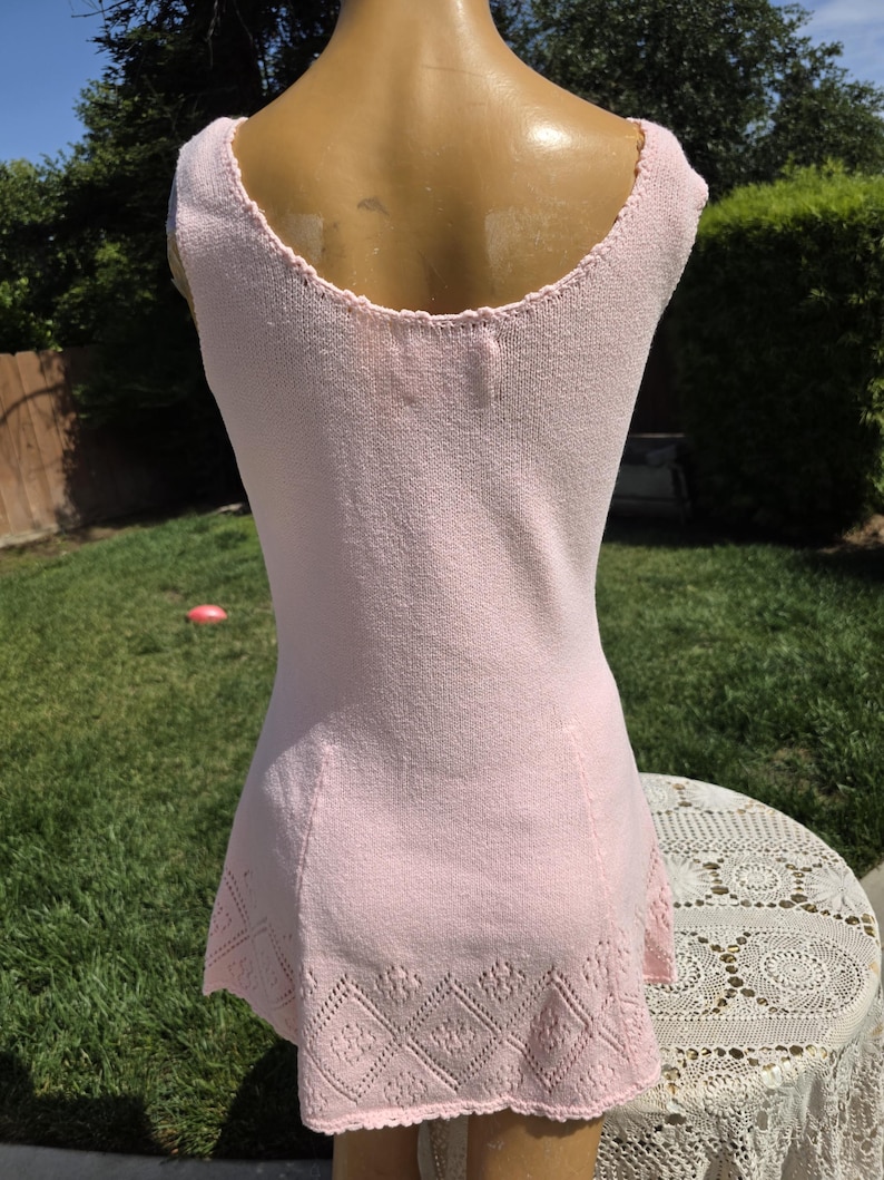 May include: A light pink sleeveless knit dress. The dress features a scoop neckline and a decorative openwork hemline. The dress is displayed on a mannequin outdoors, with a green lawn and a white lace-covered table in the background.
