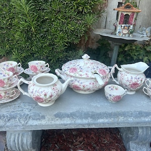 Könnte beinhalten: Ein Vintage-Porzellan-Teeservice mit floralem Muster. Das Set umfasst eine Teekanne, eine Zuckerdose, einen Sahnekännchen, eine Terrine sowie mehrere Tassen und Untertassen. Die Teile sind weiß mit rosa und roten Rosen.