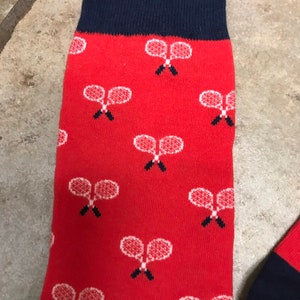 May include: Red socks with a navy blue cuff and toe. The socks feature a pattern of white tennis rackets. The socks are made of a soft, comfortable material.
