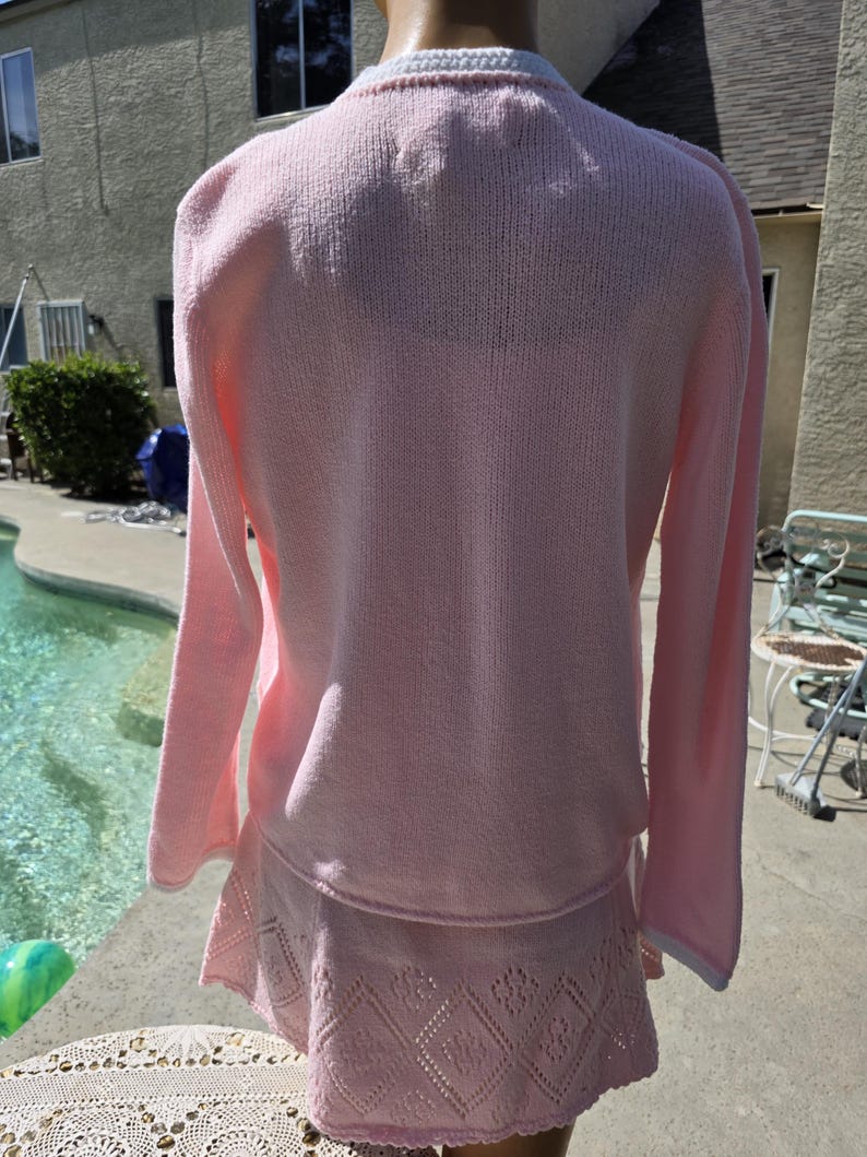 May include: A light pink knit sweater and skirt set. The sweater has long sleeves and a white trim around the collar and cuffs. The skirt has a decorative openwork pattern around the hem.