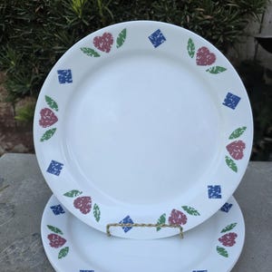 May include: White ceramic plates with a decorative border featuring red heart-shaped designs, blue squares, and green leaves. The plates are stacked, showcasing the pattern. The plates are on a stone surface.