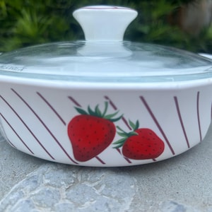 Può includere: Pirofila in ceramica bianca con coperchio in vetro. Il piatto presenta un disegno di fragole con bacche rosse e foglie verdi. Strisce diagonali bordeaux adornano i lati. Il coperchio ha un pomello bianco.