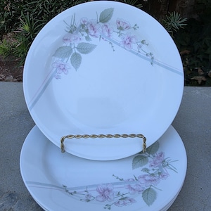May include: A stack of white ceramic plates with a floral design. Each plate features a delicate pattern of pink flowers and green leaves, accented by a gray stripe. The plates are stacked on a gold-colored metal plate holder.