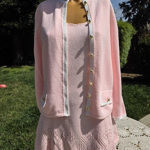 May include: A light pink knit dress set, including a cardigan and a sleeveless dress. The cardigan has gold buttons and white trim. The dress features a diamond pattern at the hem. The set is displayed on a mannequin outdoors.