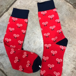 May include: A pair of red socks with navy blue cuffs, heels, and toes. The socks feature a pattern of white tennis rackets. These socks are a fun accessory for sports fans.