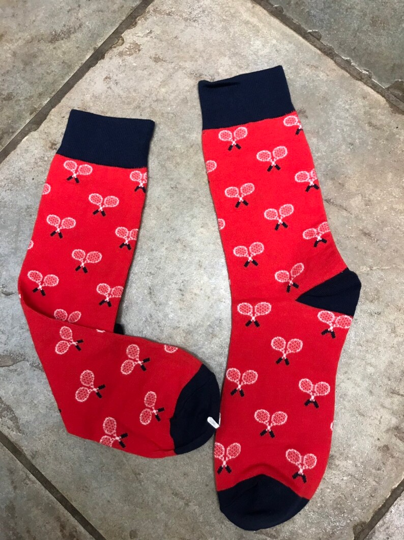 May include: A pair of red socks with navy blue cuffs, heels, and toes. The socks feature a pattern of white tennis rackets. These socks are a fun accessory for sports fans.