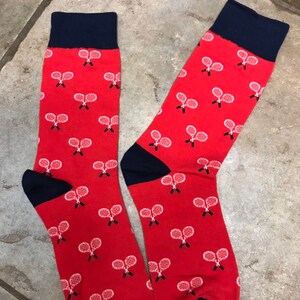 May include: A pair of red socks with navy blue cuffs and toes. The socks feature a pattern of white tennis rackets. These socks are a fun accessory for sports fans.