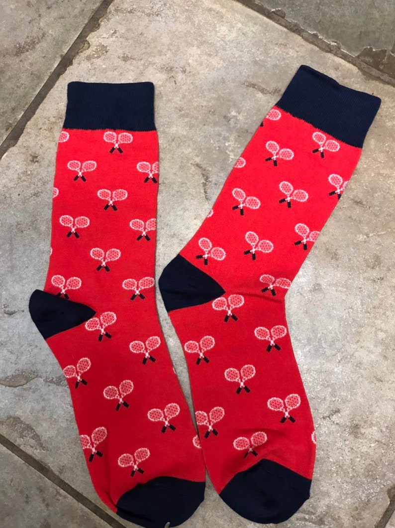 May include: A pair of red socks with navy blue cuffs and toes. The socks feature a pattern of white tennis rackets. These socks are a fun accessory for sports fans.