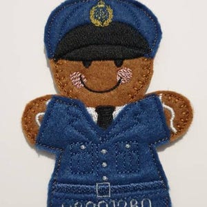 May include: A felt fridge magnet in the shape of a gingerbread man dressed as a police officer. The gingerbread man has a brown face, arms, and legs, and is wearing a blue uniform with the number R8201280. The magnet has a black hat and tie.