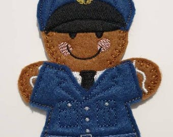 May include: A felt fridge magnet shaped like a gingerbread man dressed as a police officer. The gingerbread man has a brown face, arms, and legs, and is wearing a blue uniform with the number R8201280. The magnet has a black hat and tie.