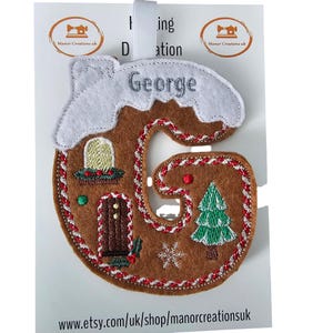 May include: A felt Christmas decoration shaped like the letter 'G', designed as a gingerbread house. It features a white snowy roof, a green Christmas tree, and the name 'George' embroidered in silver. The ornament has a red stitched border.