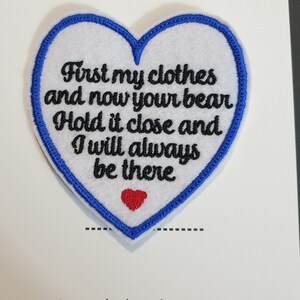 Op de afbeelding: Witte, hartvormige geborduurde patch met blauwe stiksels rond de randen. De patch heeft de tekst "First my clothes and now your bear Hold it close and I will always be there" in zwarte borduurwerk. Een klein rood hart is geborduurd in het midden onderaan de patch.