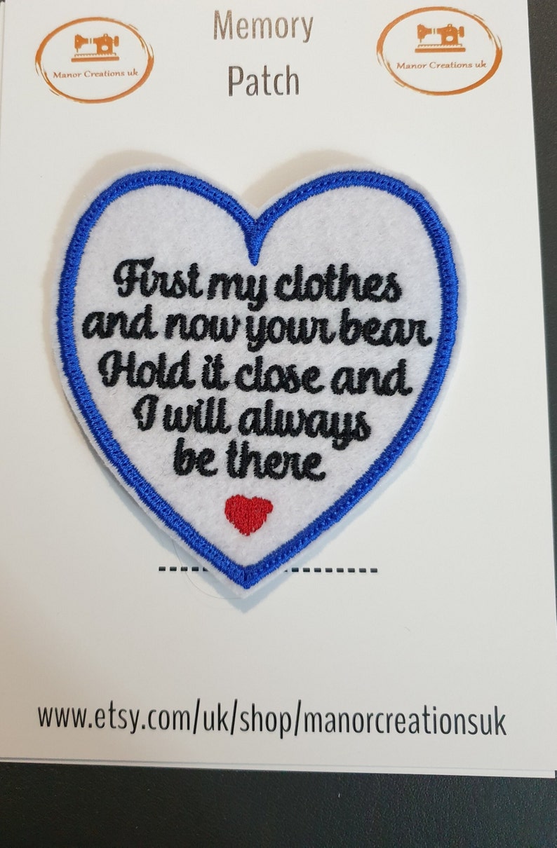 Op de afbeelding: Witte, hartvormige geborduurde patch met blauwe stiksels rond de randen. De patch heeft de tekst "First my clothes and now your bear Hold it close and I will always be there" in zwarte borduurwerk. Een klein rood hart is geborduurd in het midden onderaan de patch.